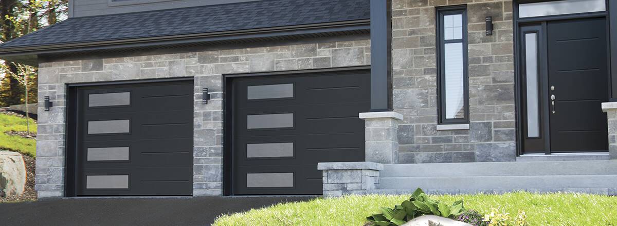 Portes de garage contemporaine noire modèle Vog avec fenêtres Harmony à gauche sur une maison grise en pierre